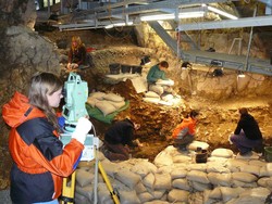 Aufnahme einer Felshöhle. Vier Menschen sitzen auf dem sandigen Höhlenboden und arbeiten an Funden. Im Vordergrund ist eine kniehohe Mauer aus weißen Sandsecken aufgebaut. Hinter dieser Mauer steht eine junge Frau in Funktionsjacke. Sie dokumentiert die Ausgrabung mithilfe eines Messgeräts.