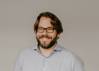 Porträtfoto von Marius Busemeyer in Frontalansicht. Zu sehen ist ein lächelnder Mann mit Vollbart und Brille bis zur Brust. Er trägt dunkelblondes volles Haar.