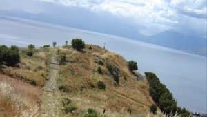 Steinweg auf einem kargen, grasbewachsenen Hügel, der zum Meer führt. An der Klippe steht ein Kreuz.
