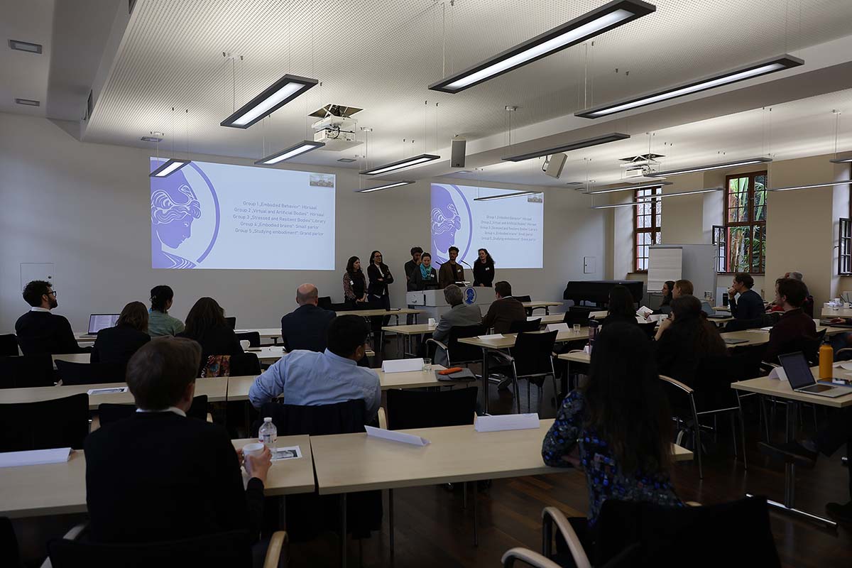 Ein Konferenzraum. Im Vordergrund sitzen in mehreren Reihen Personen. Im Hintergrund eine Gruppe an einem Rednerpult, die Wand zeigt eine Präsentation.