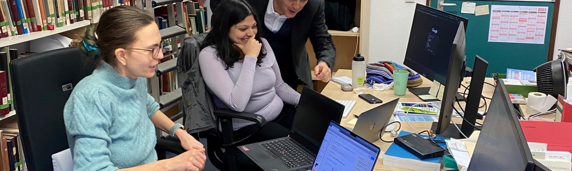 drei Personen in einem Büro in Seitenansicht. Zwei Frauen sitzen vor Laptops an einem Schreibtisch, ein Mann steht daneben und zeigt auf den Bildschirm, den alle drei interessiert anschauen. Am linken Bildrand eine Bücherregal, rechts ist der Schreibtisch mit weiteren Bildschirmen zu sehen.
