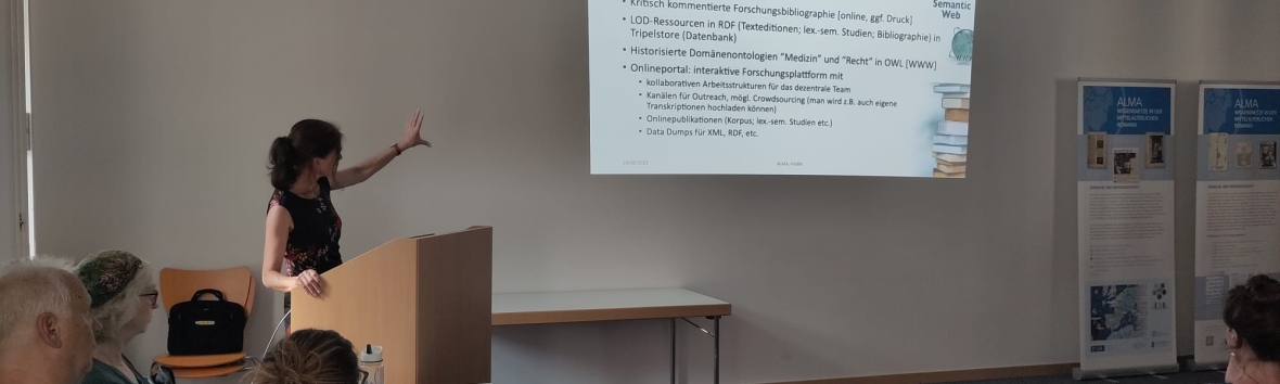 Sabine Tittel steht hinter einem Redepult und weist mit einer Hand auf die PowerPoint-Präsentation, die an der dahinterliegenden Wand angezeigt wird. Im Vordergrund sind einige Mitglieder des Teams der Forschungsstelle "Bibelglossare" zu sehen.