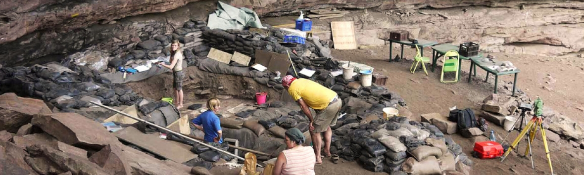 Aufnahme einer Ausgrabung. Vor einer Felswand sind Sandsäcke aufgetürmt sowie Messgeräte und Tische aufgestellt. In der Szene stehen vier Personen und kommunizieren über ihre Arbeit.