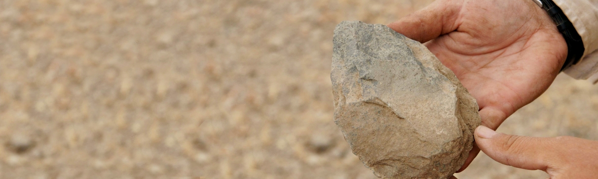 zwei Händehalten einen archäologischen Fund, ein Steinwerkzeug, vor sandfarbenem Grund in die Kamera