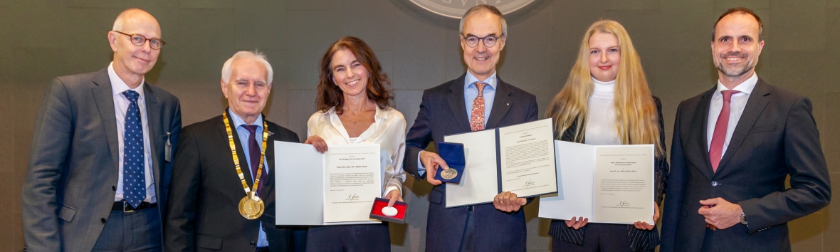 Sechs Personen stehen vor einer dunkelgrau gekachelten Wand. Die mittleren drei Personen, zwei Frauen und ein Mann, halten jeweils eine Urkunde und eine Medaille in der Hand.