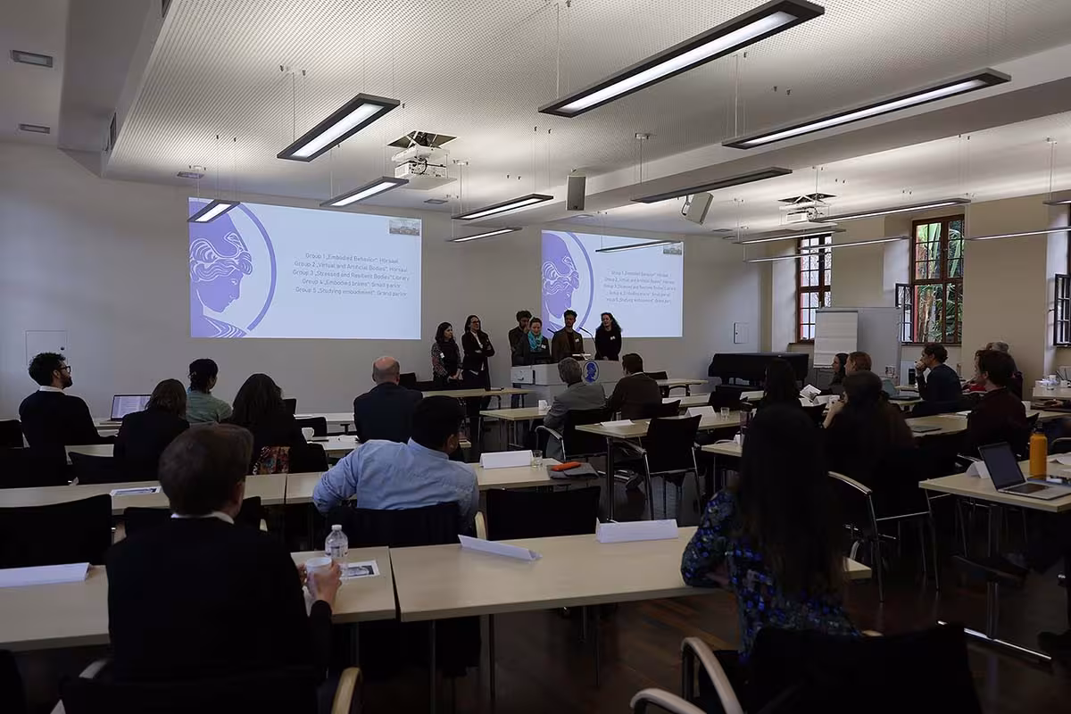 Ein Konferenzraum. Im Vordergrund sitzen in mehreren Reihen Personen. Im Hintergrund eine Gruppe an einem Rednerpult, die Wand zeigt eine Präsentation.