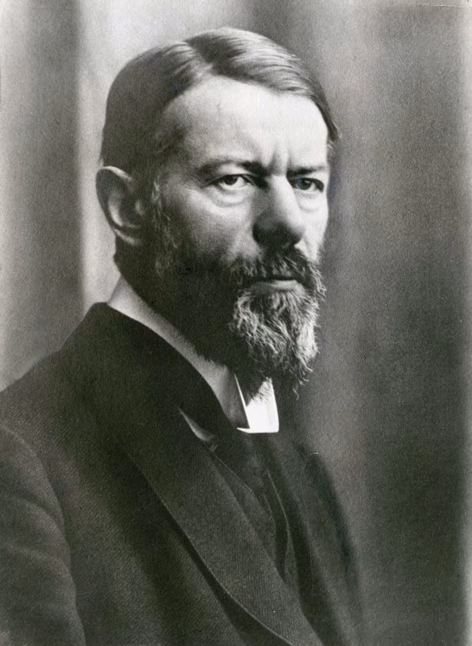 Schwarzweiß-Foto eines Mannes mit längerem Vollbart im Dreiviertelprofil. Er trägt einen Seitenscheitel und ist mit einem Anzug bekleidet. Es ist Max Weber.