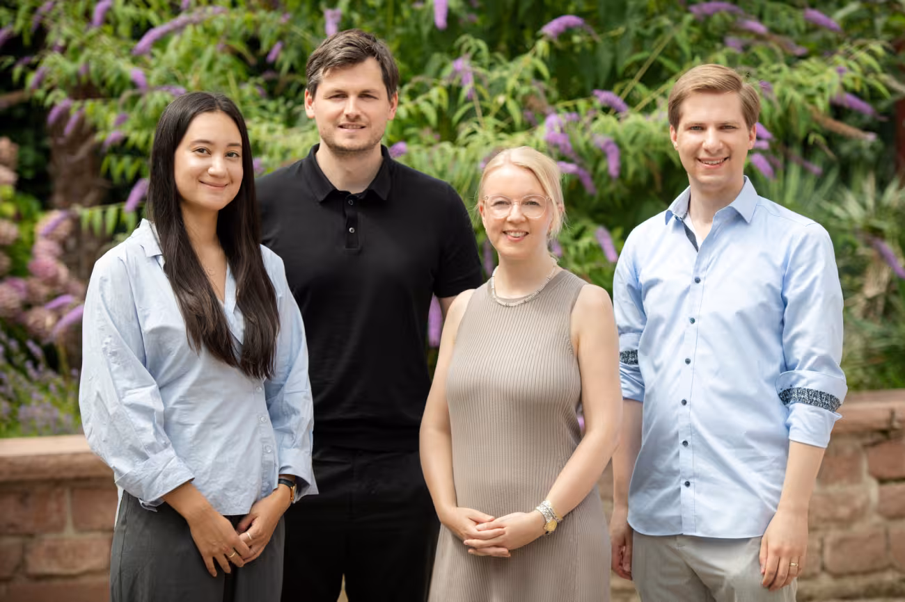 Aufnahme des Projektteams „Polykrisen“ im Garten der Akademie. Die Teammitglieder sind in der Halb-Totale zu sehen. Etwa hüfthoch im Hintergrund eine Mauer, dahinter im Hintergrund blühender Garten. Von links nach rechts im Vordergrund Stella Kim, Gordon Friedrichs, Natalie Rauscher und Steven Schwarz