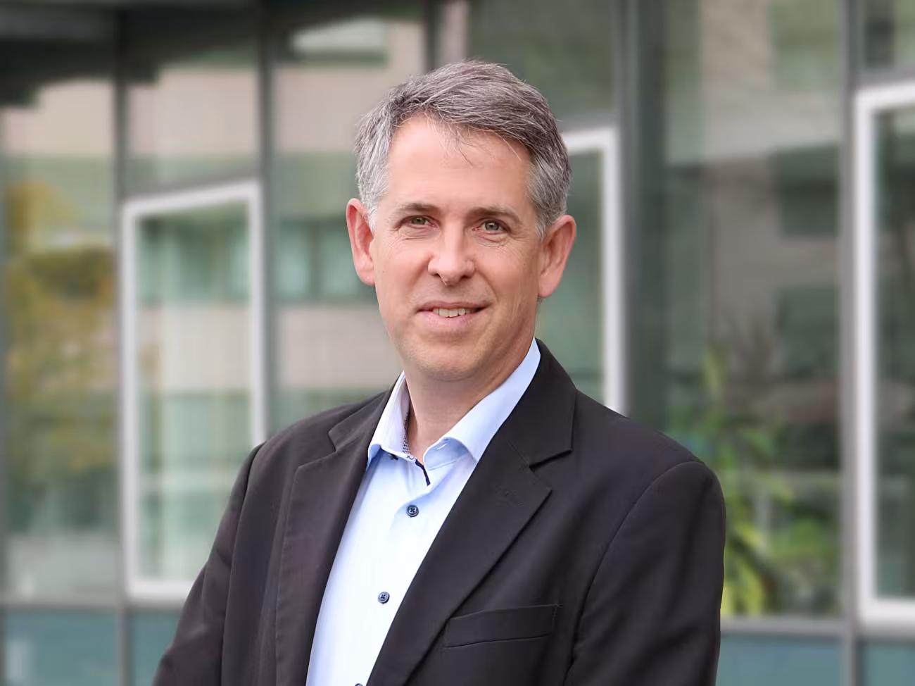Stefan Pfister, ein Mann mit grauen kurzen Haaren, die zum Seitenscheitel gekämmt sind, lächelt in die Kamera. Er trägt ein helles Hemd und darüber ein dunkles Sakko.ein 