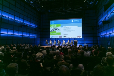 Publikumsfoto der Konferenz "Autonomes Fahren". Das Foto ist aus den hinteren Reihen des Publikums aufgenommen und Richtung Bühne fotografiert. In dem abgedunkelten Saal, der von blauen Lichtern an der Seite beleuchtet wird sitzt das Publikum und schaut zu einer Podiumsdiskussion auf der Bühne hinauf. Hinter den Diskutant*innen wird eine PowerPoint angestrahlt.