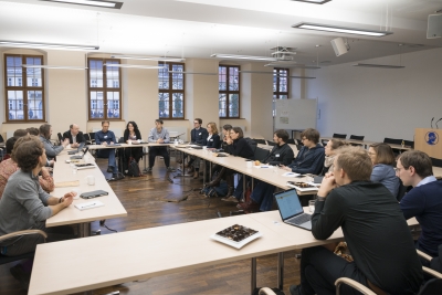 Aufnahme von einer Konferenz im Hörsaal der Akademie, eine Gruppe von etwa 20 Personen sitzt um im Viereck aufgestellte Tischreihen mit dem Gesicht in die Mitte gerichtet. Einige haben Schreibutensilien, andere Laptops vor sich aufgestellt. Im Hintergrund ist eine Fensterreihe mit fünf holzgerahmten Fenstern zu sehen.