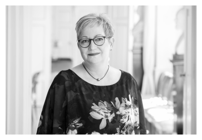 Schwarz-weiß Portrait in halbnaher Aufnahme. Frau Fuchs lächelt mit leicht zur Seite geneigtem Kopf in die Kamera. Sie trägt eine dunkle Bluse mit Blumenmuster. Dazu eine dunkel gerahmte Brille und einen Kurzhaarschnitt. Im Hintergrund sieht man den Großen Salon auf der Bel Etage der Akademie.