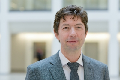 Frontalansicht bis zur Brust von Christian Drosten, der den Betrachter freundlich anblickt. Er trägt einen Anzug mit Krawatte und hat kurze dunkelbraune Haare. 