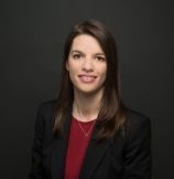 Portrait einer braunhaarigen Frau vor schwarzem Hintergrund. Sie trägt einen schwarzen Blazer und ein rotes Oberteil.