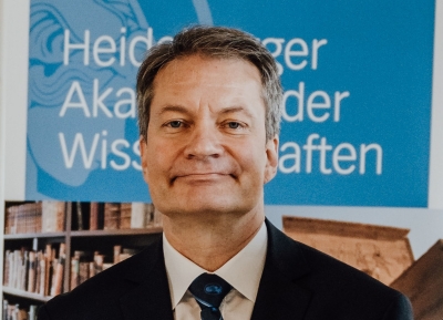 Frontalansicht bis zur Brust von Volker Wulfmeyer. Er trägt kurze Haare, grau-meliert, mit einem Seitenscheitel und lächelt dem Betrachter zu. Er steht vor einem Roll-up mit der Aufschrift "Heidelberger Akademie der Wissenschaften"