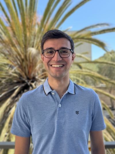 Portrait eines braunhaarige Mannes mit Brille vor einer Palme. Er trägt ein hellblaues Polo-Hemd.