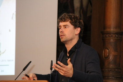 Johannes Krause spricht in der Alten Aula der Uni Heidelberg. Es ist ein farbiges Dreiviertelporträt. Er hat dunkle Locken und ein dunkles Hemd.