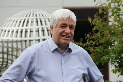 Foto von Gerhard Huisken, der den Betrachter anlächelnd bis zur Brust dargestellt ist. Das Foto zeigt ihn im Hemd mit weißen kurzen Haaren, die nach vorne gekämmt in die Stirn fallen. Im Hintergrund ist ein grüner Baum, sowie ein Pflanzengitter zu sehen. Beides steht vor einem Gebäude, dessen offenen Eingang man sieht.