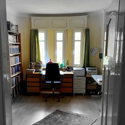 Sekretariat der Arbeitsstelle Goethe-Wörterbuch in Tübingen. Vor einem Fenster steht ein hölzerner Schreibtisch mit Schubladen links und rechts. Davor ein klassischer Schreibtischstuhl mit Rollen. Rechts vom Schreibtisch stehen zwei weitere Rollkästen mit Schubladen und technische Geräte wie ein Drucker und ein Scanner. An der linken Wand steht ein Bücherregal. Das Bild ist vom Türrahmen aus aufgenommen, der links und rechts vorne zu sehen ist.