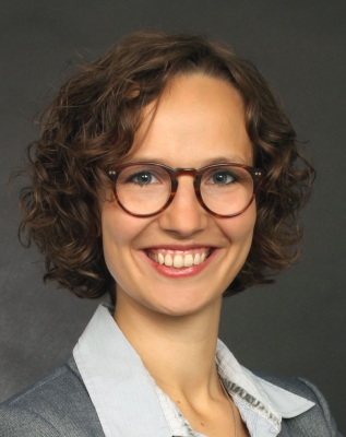 Portrait in Nahaufnahme von Paula Zschoche. Sie lächelt frontal in die Kamera. Sie trägt eine weiße Bluse und einen grauen Blazer, dazu eine dunkel gerahmte Brille. Die lockigen Haare sind kinnlang.