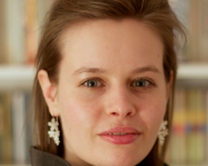 Portrait in Nahaufnahme von Helen Ahner. Sie schaut mit neutralem Blick in die Kamera. Sie trägt eine dunkle Sweatshirtjacke. Die Haare sind kinnlang, dazu trägt sie weiße Blumenohrringe. Helen Ahner ist vor einem Bücherregal abgelichtet.