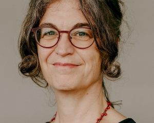 Portrait von Claudia Diehl in Nahaufnahme. Sie lächelt in die Kamera, trägt die Haare hochgesteckt und eine rotgerahmte Brille. Dazu trägt sie eine schwarze Bluse und eine Kette mit roten Perlen.