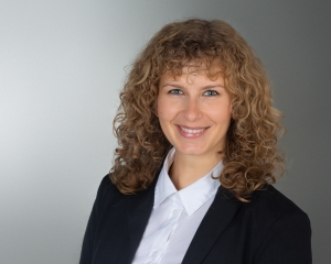 Portrait von Natalie Schunck in halbnaher Aufnahme. Sie lächelt in die Kamera. Sie trägt eine weiße Bluse und ein schwarzes Jackett. Die lockigen Haare sind schulterlang.