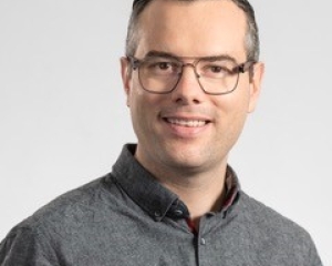 Portrait von Alexander Schlaich in halbnaher Aufnahme. Er lächelt frontal in die Kamera. Er trägt ein graues Hemd und eine dunkel gerahmte Brille. Die Haare trägt er kurz.