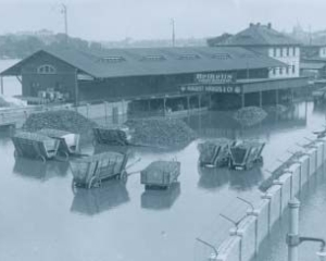 Schwarz-weiß Foto des Elbehochwassers im Januar 1932: Die Elbe tritt in der Nähe von Dresden über ihre Ufer (Bundesarchiv, Bild 102-12896, anonymes undatiertes Foto)​; zu sehen sind ein längliches Gebäude und einige Kutschwagen, deren Räder im Wasser stehen. 
