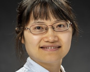 Portrait in Nahaufnahme von Jingyuan Xu. Sie lächelt in die Kamera. Sie trägt eine blau-weiß gestreifte Bluse. Die dunklen Haare sind zusammengebunden, dazu trägt sie einen Pony und eine schmal gerahmte Brille.