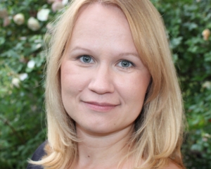 Portrait in Nahaufnahme von Mari Sepp. Ihre Schultern sind seitlich zur Kamera gedreht, sie lächelt mit dem Gesicht frontal zur Kamera geneigt. Sie trägt ein schwarzes Oberteil und ist vor einem Rosenbusch abgebildet. Die blonden Haare sind schulterlang.