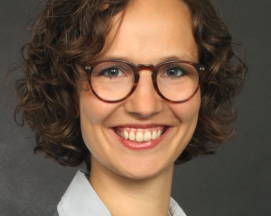 Portrait in Nahaufnahme von Paula Zschoche. Sie lächelt frontal in die Kamera. Sie trägt eine weiße Bluse und einen grauen Blazer, dazu eine dunkel gerahmte Brille. Die lockigen Haare sind kinnlang.