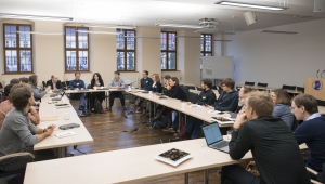 Aufnahme von einer Konferenz im Hörsaal der Akademie, eine Gruppe von etwa 20 Personen sitzt um im Viereck aufgestellte Tischreihen mit dem Gesicht in die Mitte gerichtet. Einige haben Schreibutensilien, andere Laptops vor sich aufgestellt. Im Hintergrund ist eine Fensterreihe mit fünf holzgerahmten Fenstern zu sehen.