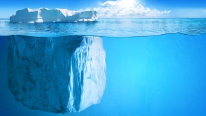 Foto eines Eisbergs im Ozean. Das Bild ist aus der Höhe der Wasseroberfläche aufgenommen und zeigt sowohl den Eisberg über Wasser als auch den sehr viel größeren Teil des Bergs unter Wasser. Am Horizont sind eine Wolkenbank und die Sonne zu sehen.