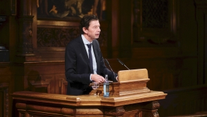 Dreiviertelporträt von Prof. Christian am Pult stehend und sprechend. Er hat dunkle lockige Haare und trägt einen Anzug mit weißem Hemd und Krawatte.
