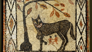Quadratisches Mosaik, das im Zentrum eine Wölfin mit zwei kleinen Personen darunter zeigt. Die Wölfin steht unter einem Baum. Am Rand verläuft ein Muster aus aneinandergereihten Rauten.