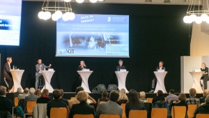 Raumaufnahme eines Vortragssaals, eine PowerPoint wird an eine schwarze Wand gestrahlt, vor der fünf zwei bezogenen Stehtische für eine Podiumsdiskussion stehen. An jedem Tisch steht eine Person, im Vordergrund des Bildes ist das Publikum mit dem Rücken zur Kamera gedreht zu sehen