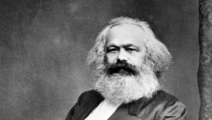 Schwarzweiß-Foto von Karl Marx. Er sitzt in einem Stuhl und blickt den Betrachter frontal an. Seine rechte Hand steckt er dabei unter das Revers seines Gehrocks, der linke Arm ruht auf der Stuhllehne.  