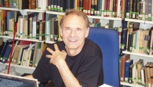 Frankwalt Möhren sitzt an einem Schreibtisch und schaut frontal in die Kamera. Neben seinem Arm liegt ein Papierstapel. Im Hintergrund ein Bücherregal.