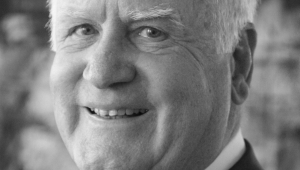 Schwarz-weiß Portrait von Manfred Fuchs in naher Aufnahme. Er hat weißes Haar und lächelt in die Kamera. Unter seinem schwarzen Jackett trägt er ein weißes Hemd sowie eine karierte Krawatte.