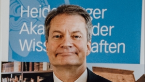 Frontalansicht bis zur Brust von Volker Wulfmeyer. Er trägt kurze Haare, grau-meliert, mit einem Seitenscheitel und lächelt dem Betrachter zu. Er steht vor einem Roll-up mit der Aufschrift "Heidelberger Akademie der Wissenschaften"