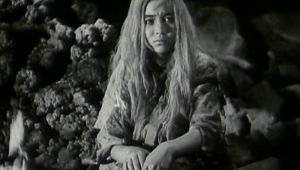 Still aus dem in schwarzweiß gedrehten chinesischen Film „Bai Mao Nü” von 1950, der das namengebende weißhaarige Mädchen sitzend in einer Höhle zeigt.