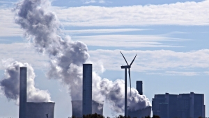 Das Bild zeigt rauchende Schloten von Fabriken, dazwischen steht ein Windrad.