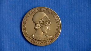 Münze mit Athene auf blauen Untergrund. Die goldene Münze zeigt den für die Akademie typischen Kopf der Athene im Profil. Um diesen herum steht "HEIDELBERGER AKADEMIE DER WISSENSCHAFTEN 1909"