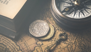 Auf einer antiken Weltkarte liegen ein antiker Schlüssel, zwei antik aussehende Münzen, ein antiker Kompass und ein dickes altes Buch mit Frakturschrift.
