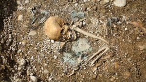 Bild eines aus dem Sandboden ragenden Skeletts. Der Kopf, die Schulter sowie ein Arm sind zu sehen. Neben dem Skelett liegen einige Schmuckstücke.