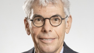 Brust-Porträt von Stefan Maul. Er trägt eine runde Brille hat kurze graue Haare und lächelt den Betrachter/die Betrachterin an. Er trägt ein Hemd, Krawatte und eine Anzugsjacke.