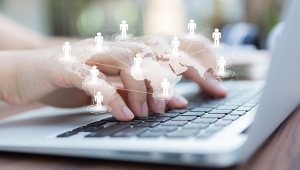 Das Bild zeigt Hände über einer Laptoptastatur. Im Vordergrund ist über den Händen eine virtuelle Weltkarte mit Personen, die auf den einzelnen Kontinenten stehen, zu sehen.