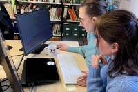Das Bild zeigt zwei Personen von der Seitenansicht an einem Schreibtisch vor einem Computerbildschirm sitzend. Eine Person hat eine Hand an der Maus, die andere schaut interssiert auf den Bildschirm. Auf dem Scheribtisch steht eine Tasse Tee und die Blätter einer Zimmerpflanze ragen vom oberen Bildrand in das Foto hinein. Im Hintergrund ein Bücherregal.