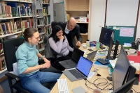 drei Personen in einem Büro in Seitenansicht. Zwei Frauen sitzen vor Laptops an einem Schreibtisch, ein Mann steht daneben und zeigt auf den Bildschirm, den alle drei interessiert anschauen. Am linken Bildrand eine Bücherregal, rechts ist der Schreibtisch mit weiteren Bildschirmen zu sehen.